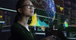 Stock photo of a young Asian woman looking at see through global & environmental data whilst seated in a dark office. The data is projected on a see through (see-thru) display.